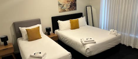 Habitación de hotel con dos camas, cada una con ropa blanca y una sola almohada marrón. Hay un cuadro en la pared, una mesita de noche con lámpara y una ventana con cortinas blancas.