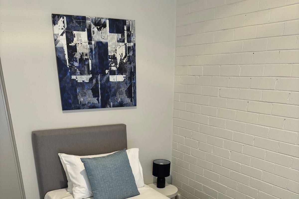 Un lit bien fait, avec une literie blanche et une tête de lit grise, une petite table de chevet avec une lampe et un tableau abstrait accroché au mur.