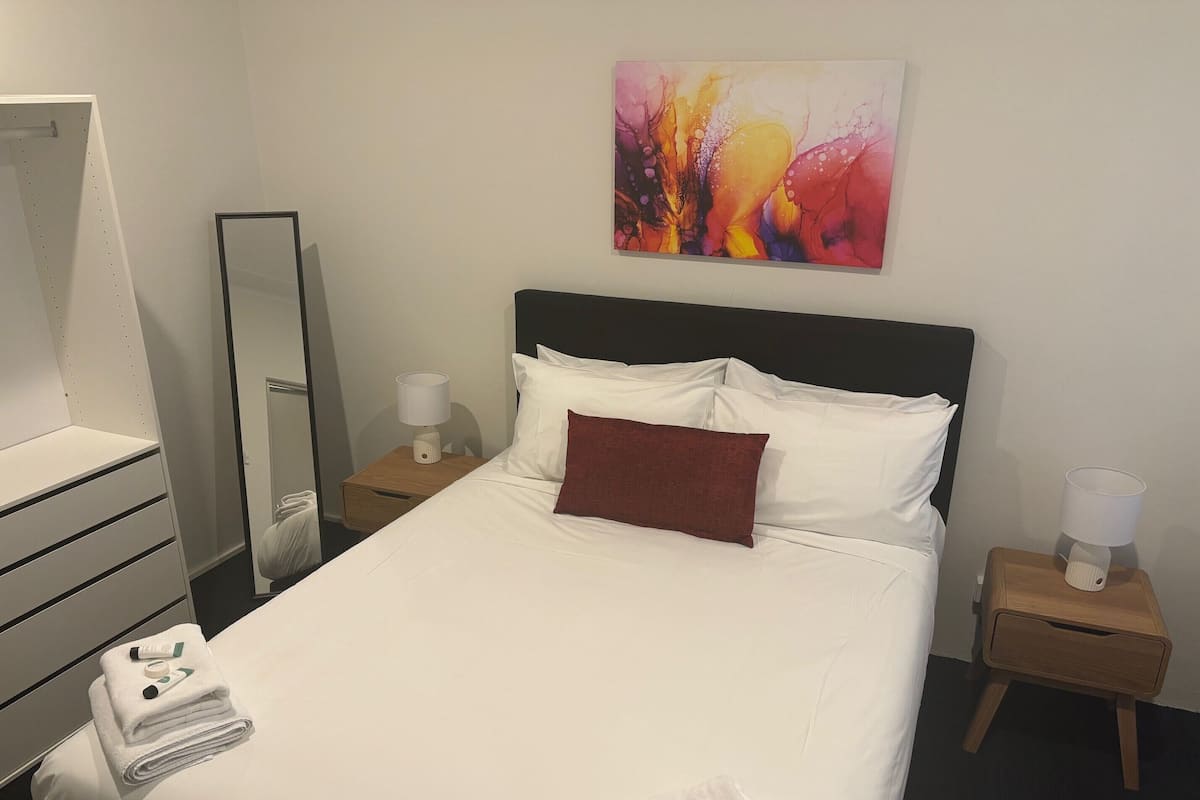 Un lit bien fait, avec du linge de lit blanc, un oreiller rouge et une serviette pliée, sur laquelle se trouvent une bouteille et un tube. Il y a une table de chevet en bois avec une lampe, ainsi qu’une petite table avec une lampe. Un grand miroir est accroché au mur, au-dessus du lit.