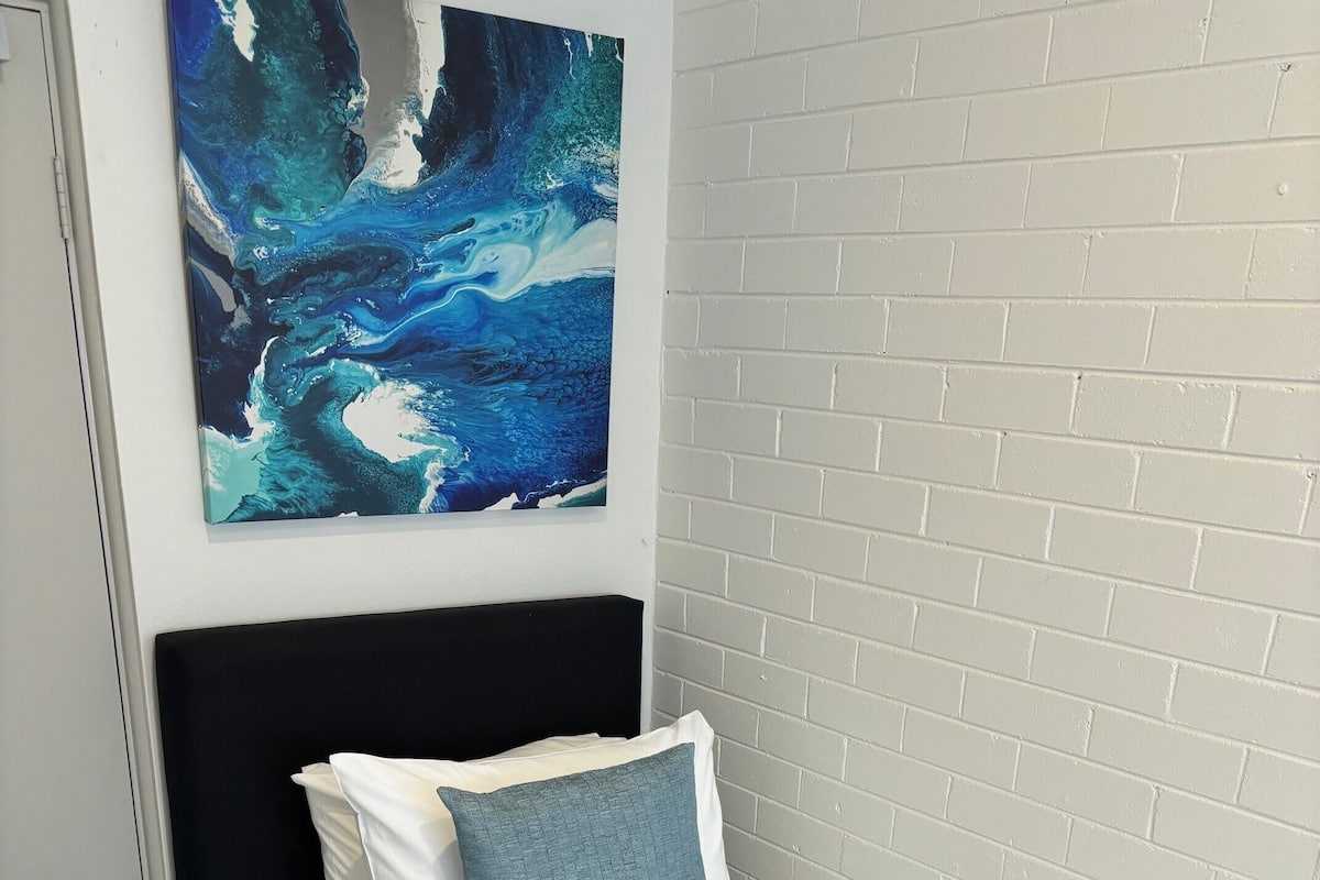 Un lit simple avec une literie blanche et un oreiller bleu, une serviette blanche et une bouteille de lotion posée à côté.