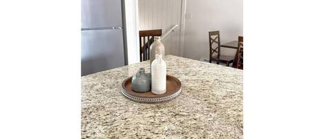 Uma bancada de cozinha com superfície de granito, uma geladeira de aço inoxidável e uma área de jantar com cadeiras e mesa de madeira.