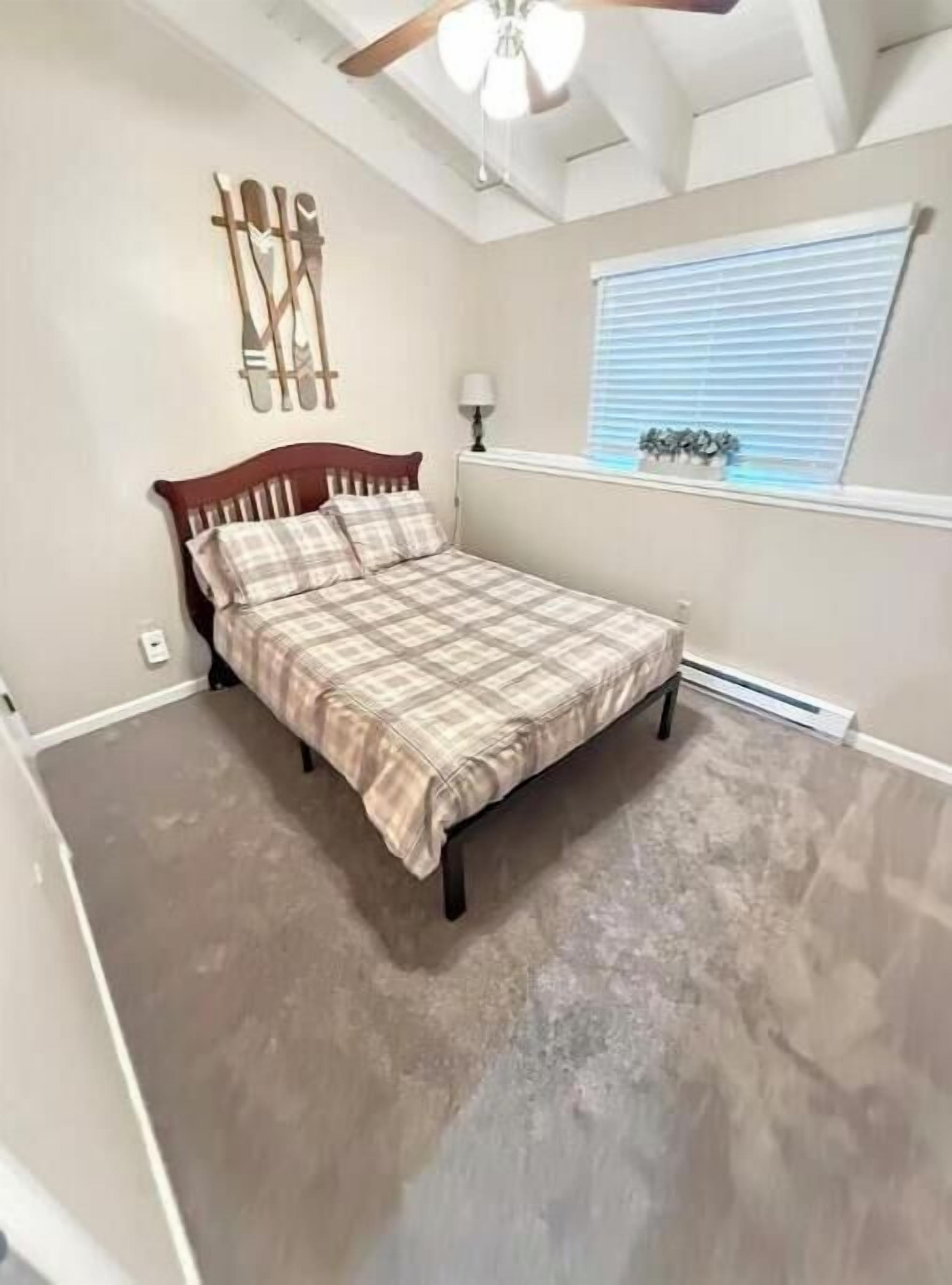 Un dormitorio con cama, ventilador de techo, ventana con persianas y una lámpara fijada a la pared.