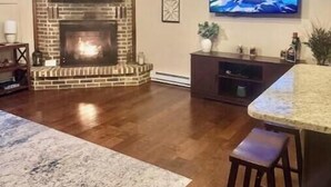 A living room with a brick fireplace, a flat-screen TV mounted on the wall, a wooden floor, and a ceiling fan.