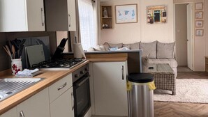 A compact kitchen with a stove, oven, and a small refrigerator. There is a wicker basket and a trash bin on the floor. A sofa and a small table are visible in the background. - Shorefield Country Park, 3 Bed,passes Included! (Lymington)