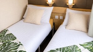 Dos camas individuales con ropa de cama blanca y mantas decorativas con estampado de hojas verdes, cabeceros de madera y luces de lectura fijadas a la pared.