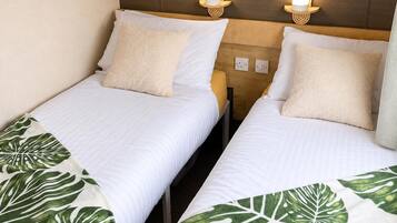 Two single beds with white bedding and green leaf-patterned throw blankets, wooden headboards, and wall-mounted reading lights.