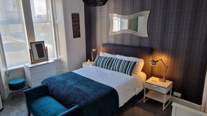 A neatly arranged bedroom with a bed, bedside tables, a chair, a window with blinds, and a wall-mounted mirror. - Troon Beachcombers Apartment (Troon)