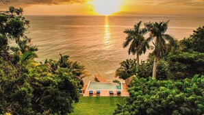 Vista a la piscina con un atardecer sobre el océano, rodeado de abundante vegetación.