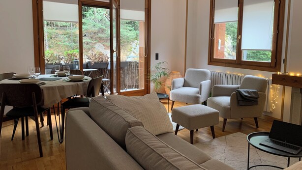 A modern living room with a sofa, chairs, a dining table set for four, and a laptop on a side table. Large windows offer a view of a garden.
