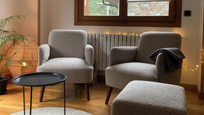 A modern living room with two grey armchairs, a small black table, and a round white rug. A large window with wooden frames and blinds is above a radiator.