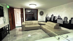 Un soggiorno con un divano angolare beige, un tavolino in vetro, un adesivo murale con vista sulla città, un televisore e una porta con la scritta 'SALIDA'.