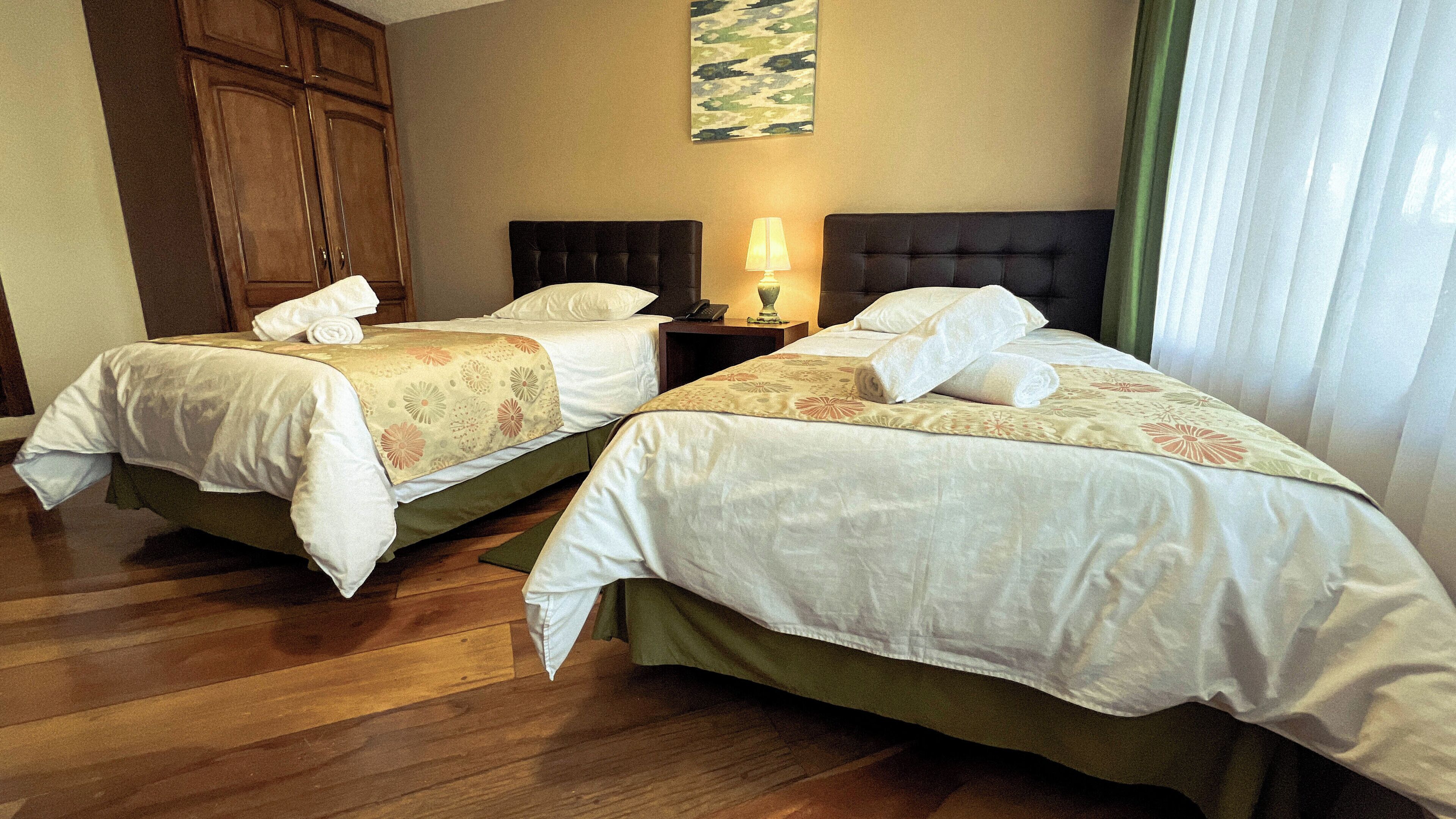 A hotel room with two beds, wooden flooring, a nightstand with a lamp, and a large painting on the wall.
