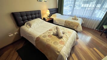 A hotel room with two beds, a nightstand with a lamp, and a view of the outdoors through sheer curtains.