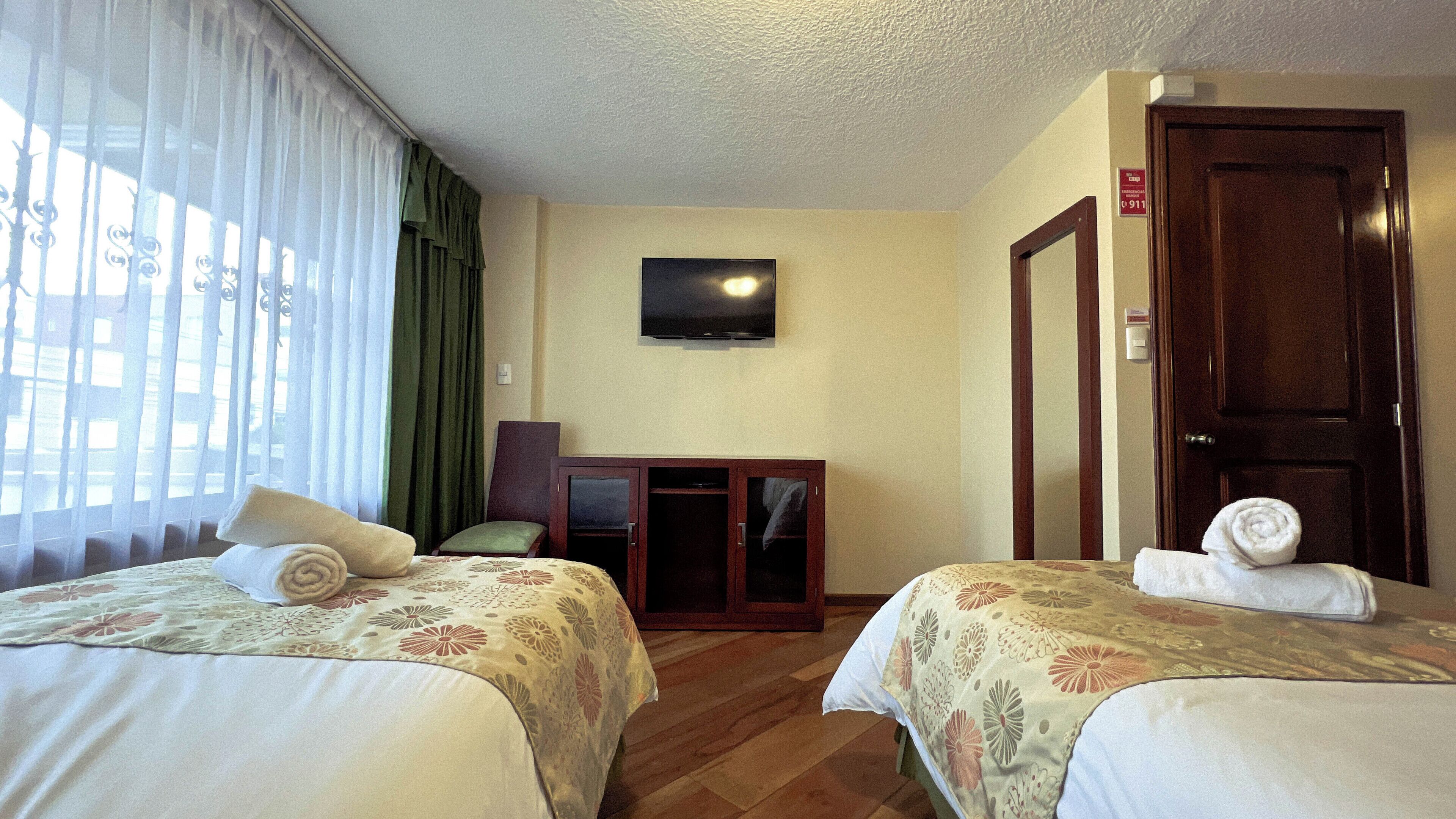 A hotel room with two beds, a television on the wall, and a door with an emergency exit sign.