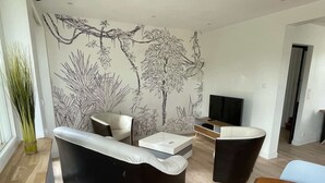 Un soggiorno moderno con un divano nero, un divano bianco e un tavolino. C'è una TV su un mobile in legno e un grande murale raffigurante alberi e piante.