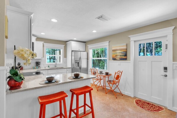 A modern kitchen with white cabinets, a stainless steel refrigerator, a dining area with orange chairs, and a red rug.
