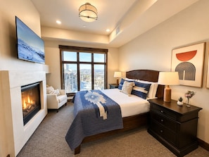 Une chambre d’hôtel avec une cheminée, un grand lit recouvert d’une couette bleue, une table de chevet avec une lampe, une commode et une grande fenêtre donnant sur la ville.