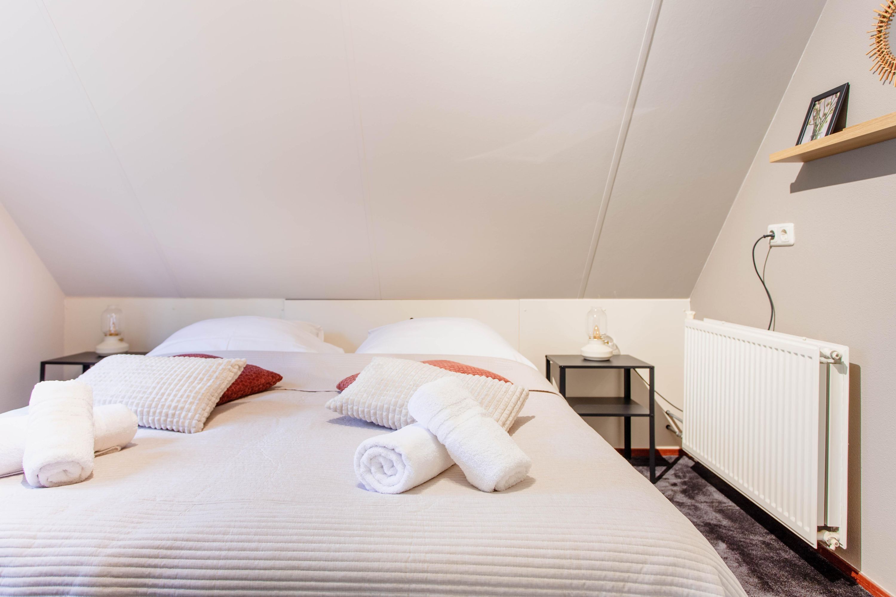 Ein ordentlich gemachtes Bett mit weißen Bettwäsche und zusammengerollten Handtüchern, ein weißes Kopfkissen, ein rotes Kopfkissen und eine kleine Ablagefläche mit Bilderrahmen an der Wand.