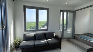 A modern living room with a grey sofa, large windows, and a view of greenery.