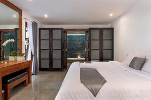 Un dormitorio moderno con una cama grande, un escritorio de madera y vistas al mar a través de puertas abiertas.