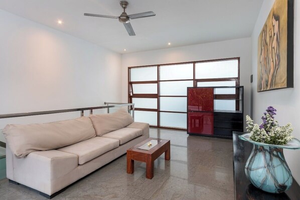 Una sala de estar moderna con un sofá color beige, una mesa de centro de madera, un florero de vidrio con flores, un ventilador de techo y un ventanal con paneles de vidrio esmerilado.