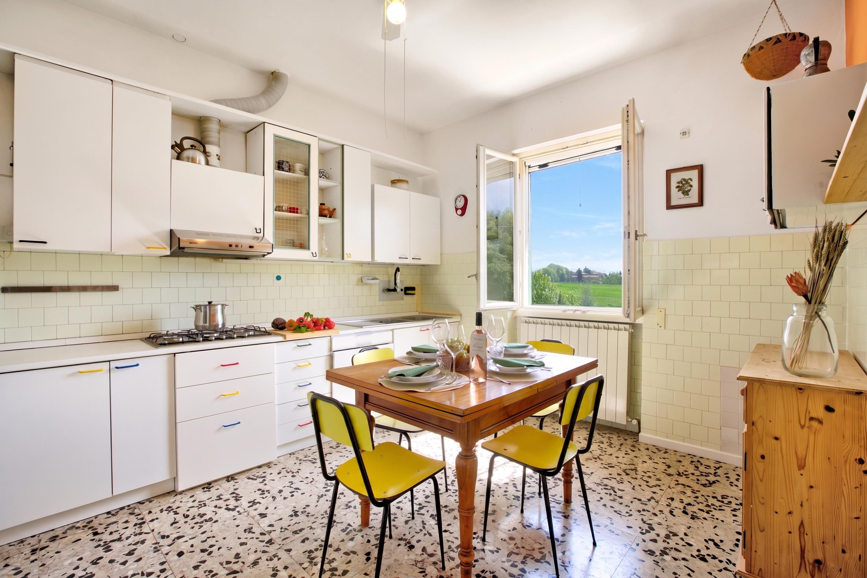 A kitchen with white cabinets, a dining table with yellow chairs, a window with a view of greenery, and a radiator under the window.