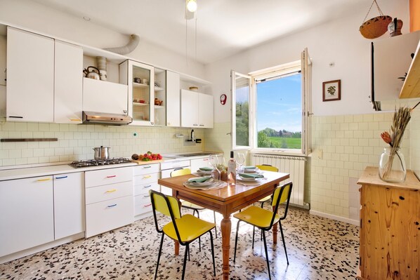 Una cocina con armarios blancos, una mesa de comedor con sillas amarillas, una ventana con vistas a la vegetación y un radiador debajo de la ventana.
