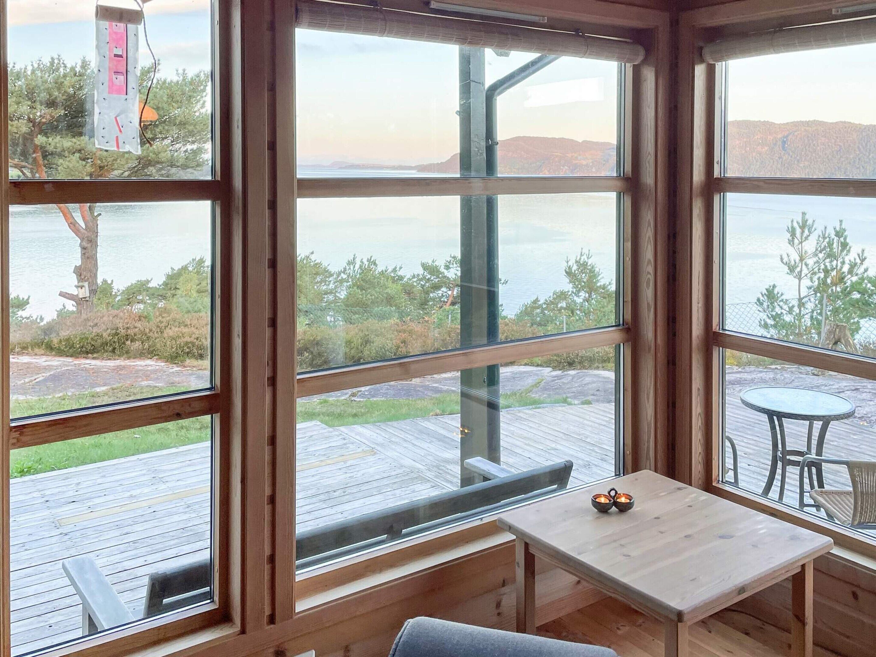 A cabin with large windows overlooking a lake, a wooden deck, and a small table with two cups.