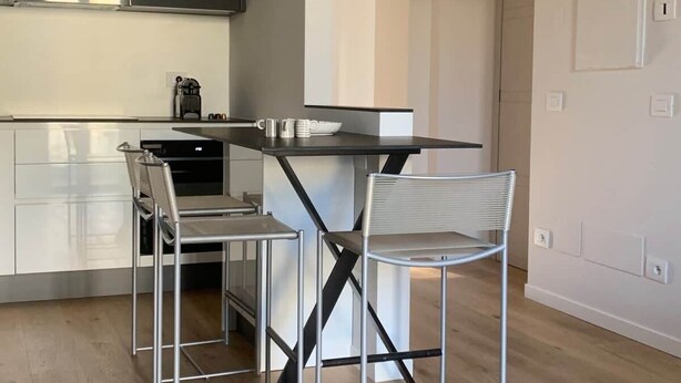 A modern kitchen with a dining area, featuring a central island, two chairs, and a coffee machine.