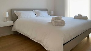 A neatly made bed with white linens and towels, a bedside table with a lamp, and a window with curtains.
