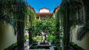 A courtyard with a central tree, surrounded by lush greenery and a building with a red roof in the background. - UDICHI REGENCY (Bolpur)