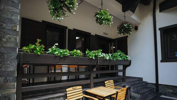 Un espace extérieur abrité, doté de bancs et de tables en bois, entouré de murs en pierre et agrémenté de jardinières suspendues remplies de verdure.