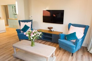 Een woonkamer met twee blauwe fauteuils, een houten salontafel en een televisie aan de muur.