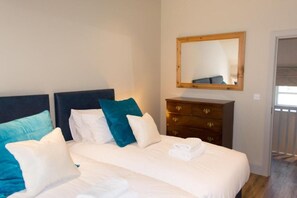 Een tweepersoonsbed met witte en teal kussens, een houten dressoir en een spiegel aan de muur.