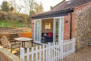 A cozy outdoor seating area with a table and chairs, a white picket fence, and a brick building with a glass door leading to a furnished room. - Host & Stay | Boathouse Barn (Frettenham)