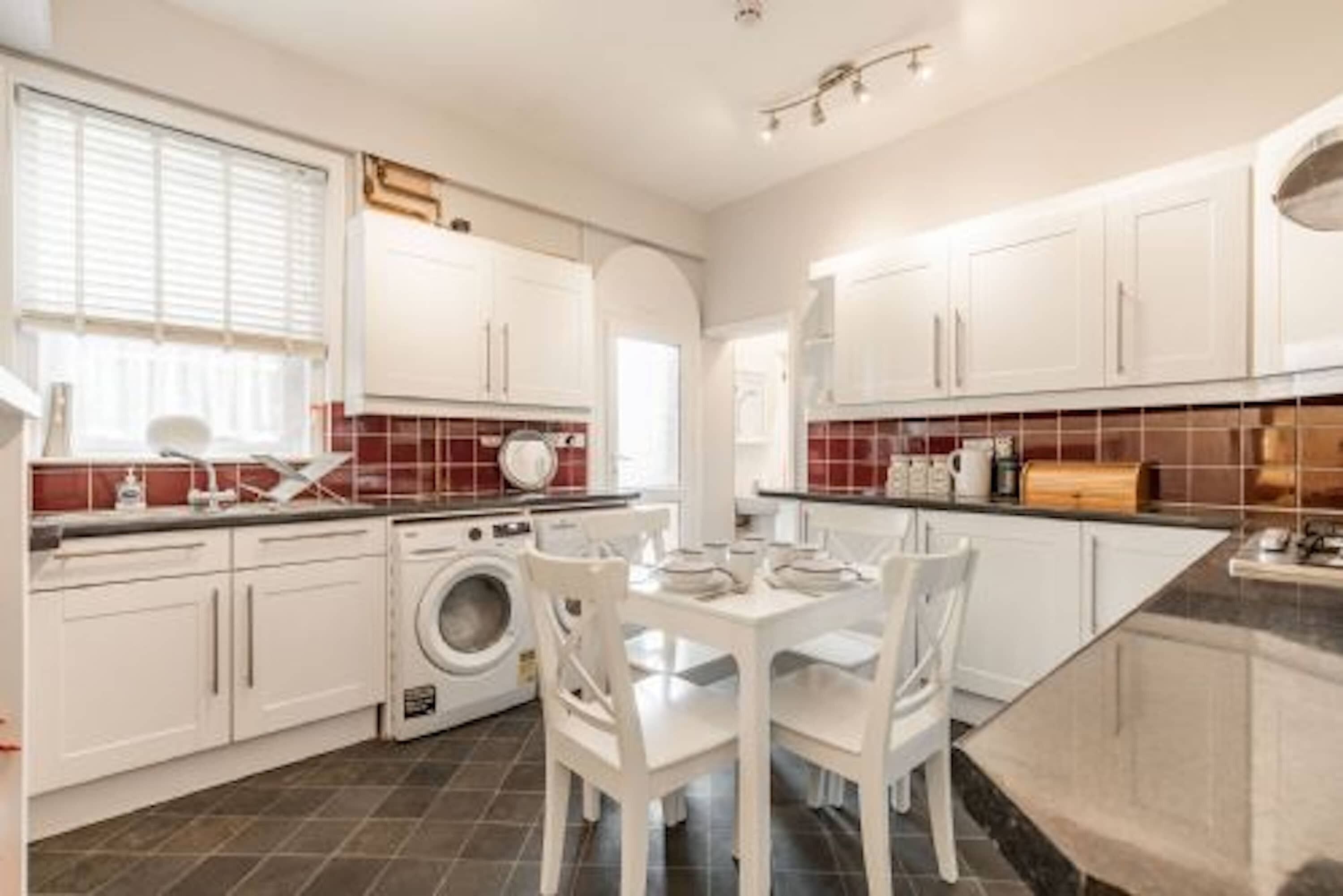 A modern kitchen with white cabinets, a washing machine, and a dining area with white chairs.