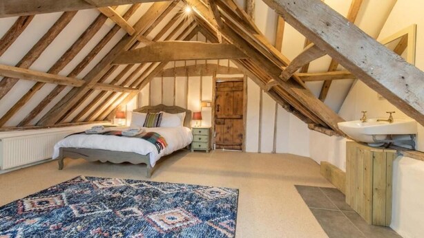 A bedroom with a sloped ceiling, a bed, a bedside table, a lamp, and a wooden cabinet with a sink. - Host & Stay | Mill House (Worthing)