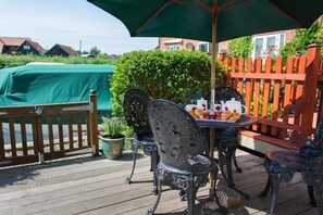 En udendørs terrasse med et bord dækket til et måltid, omgivet af metalstole og en grøn parasol.