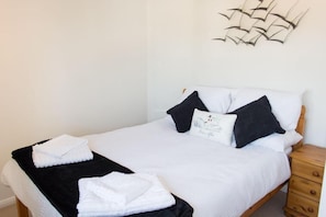 A neatly made bed with white bedding and black pillows, a wooden nightstand with drawers, and a decorative wall piece above the bed.
