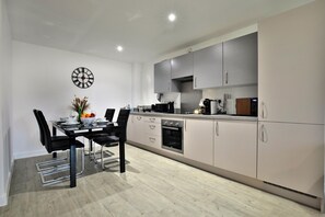 A modern kitchen with grey cabinets, a dining table with chairs, and a clock on the wall. - Manchester - 2-bed Apartment - Sleeps 4 - Wifi (Salford)
