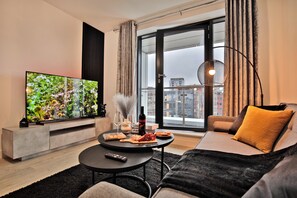 A modern living room with a large flat-screen TV, a sofa, a coffee table, and a view of the city through large windows. - Manchester - 2-bed Apartment - Sleeps 4 - Wifi (Salford)