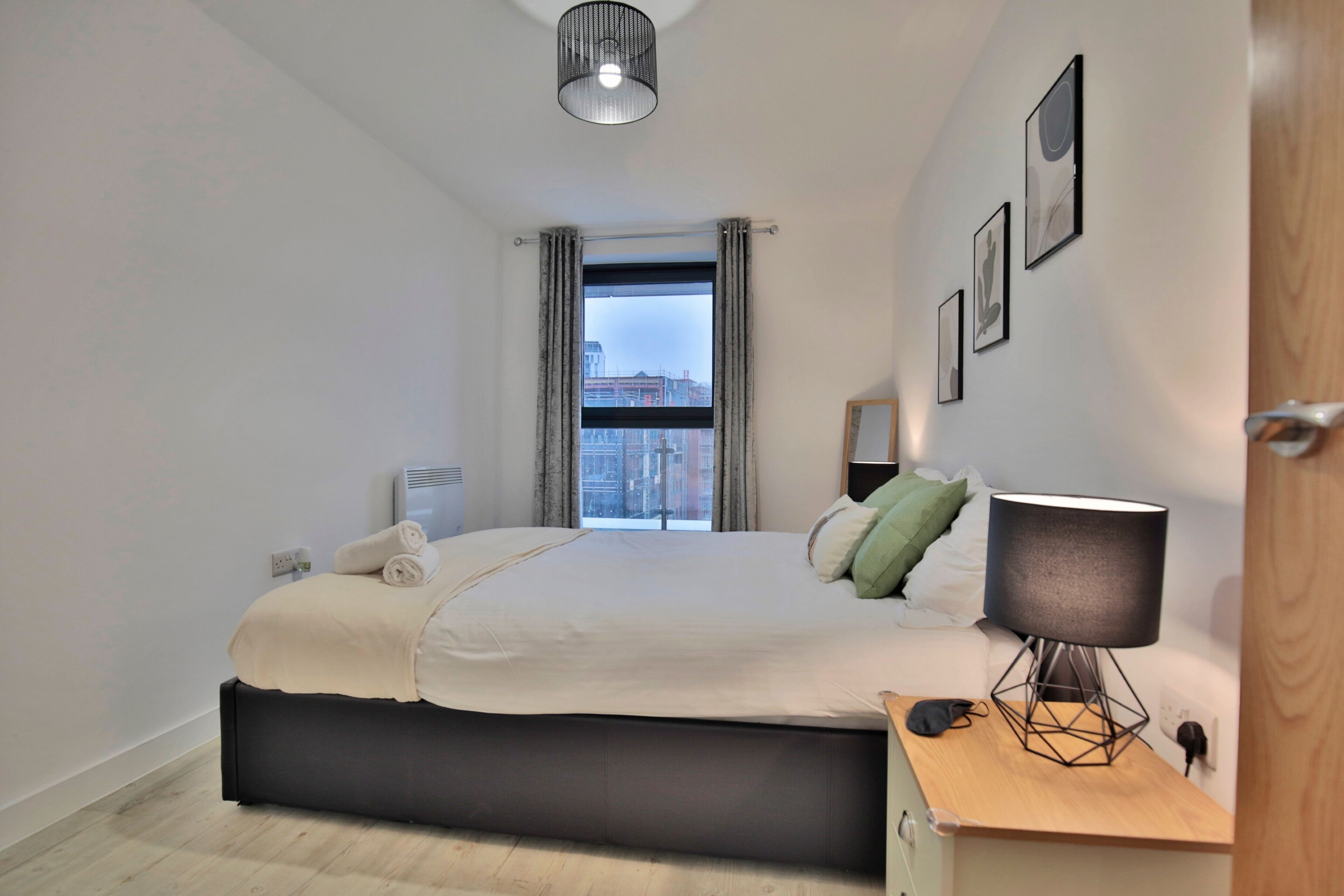 Ein modernes Hotelzimmer mit einem großen Bett, einem Nachttisch mit Lampe und Blick auf die Stadt durch das Fenster.
