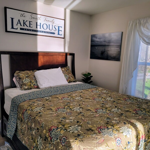 A bedroom with a bed, a floral bedspread, a headboard, a sign above the bed, a wall-mounted picture, and a window with curtains. - Sublime Sunsets--Charming Cottage with No Bad Views (King Ferry)