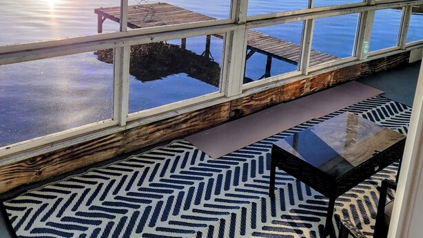 A room with large windows overlooking a calm body of water, a wooden dock, and a small table.
