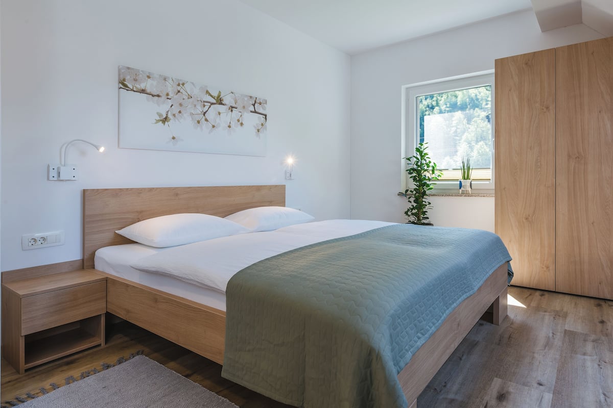 Een moderne slaapkamer met een houten bed, nachtkastjes, een nachtlampje, een raam met uitzicht op bomen, en een wandlamp.
