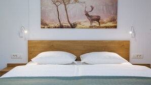 Een keurig opgemaakt bed met witte lakens en een houten hoofdbord, een aan de muur bevestigd schilderij van een hert, en bedlampjes.
