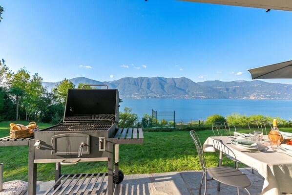 An outdoor grill setup with a table and chairs, overlooking a body of water and mountains. - Casa Antonia: Lake Maggiore Villa With Private Pool (Porto Valtravaglia)