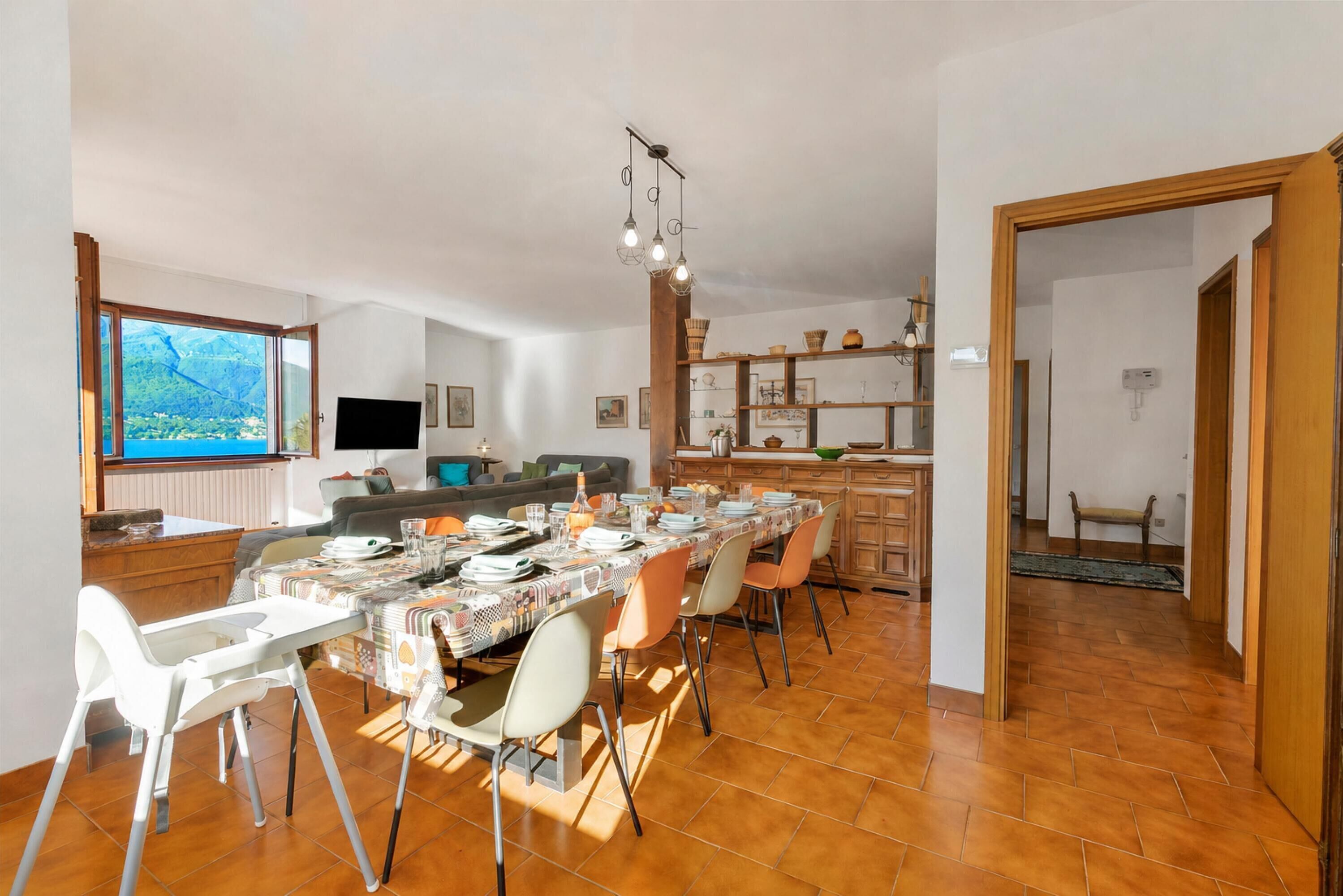 A spacious dining area with a large table set for six, a kitchen in the background, and a view of a coastal landscape through a large window.