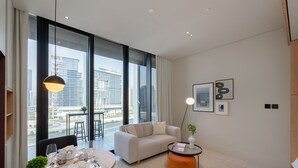 A modern hotel room with a large window, a sofa, a dining table set, and a small orange ottoman. - Cozy Studio w/ Burj View Balcony, Business Bay (Dubai)