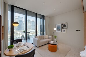 A modern hotel room with a large window, a sofa, a dining table set, and a small orange ottoman. - Cozy Studio w/ Burj View Balcony, Business Bay (Dubai)